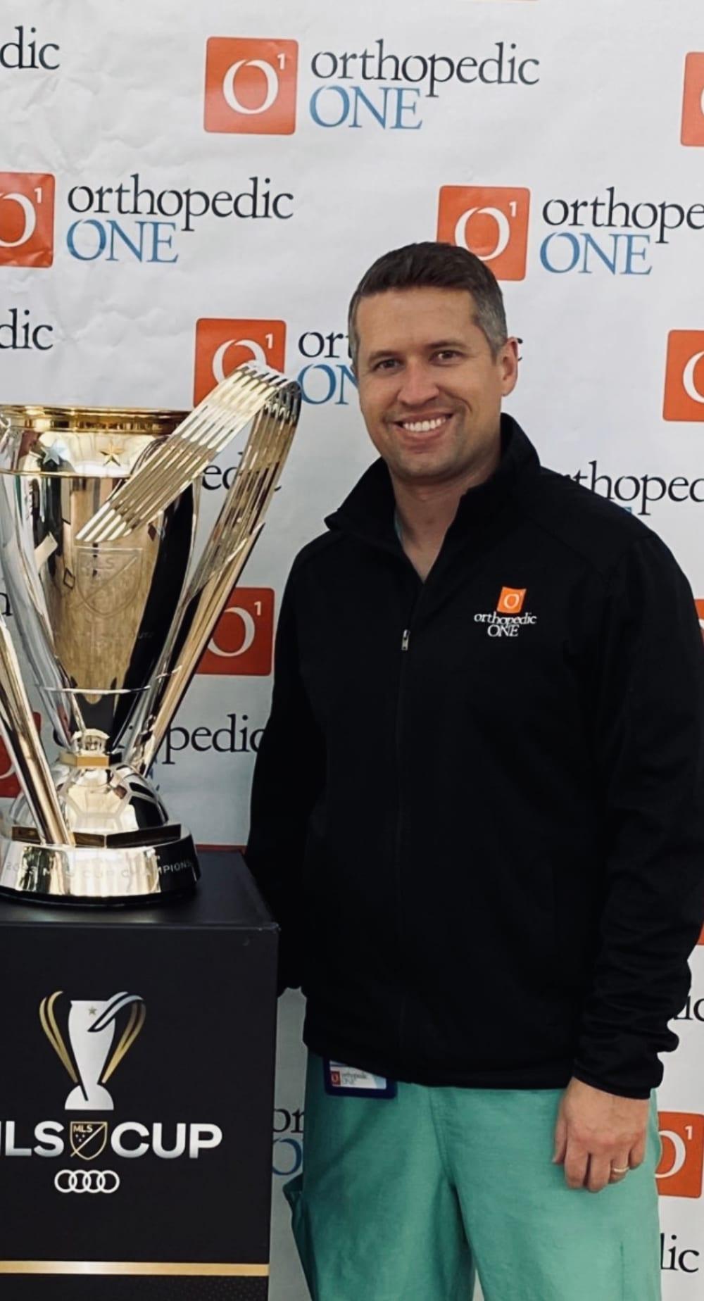 Dr. Ryan Blackwell standing beside the MLS Cup at an Orthopedic ONE event.